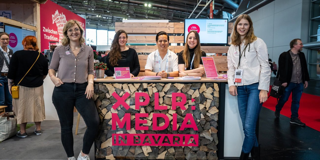 Fünf Personen stehen lächelnd an einem Messestand mit der Aufschrift 'XPLR: MEDIA in Bavaria'. Der Stand ist aus Holz und dekoriert mit Holzscheiten. Im Hintergrund sind weitere Messebesucher und ein Bildschirm zu sehen.