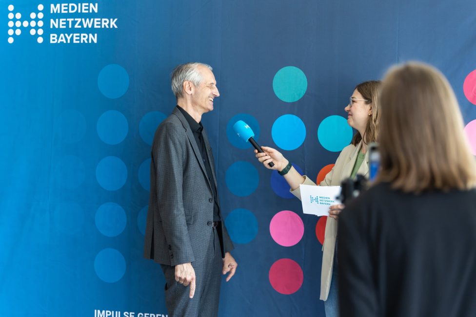 Ein Mann im Anzug wird von einer Frau mit Mikrofon interviewt. Im Hintergrund ist ein blauer Banner mit dem Logo 'Medien Netzwerk Bayern' zu sehen.