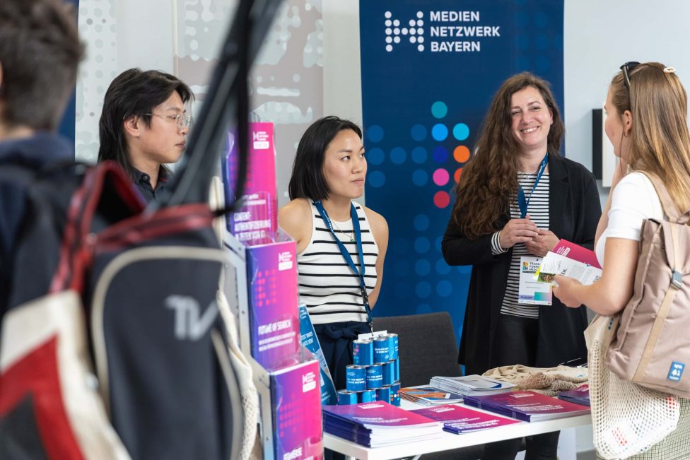 Mehrere Personen stehen an einem Informationsstand des Mediennetzwerks Bayern. Auf dem Tisch liegen Broschüren und Infomaterialien. Eine Frau lächelt, während sie mit einer Besucherin spricht.