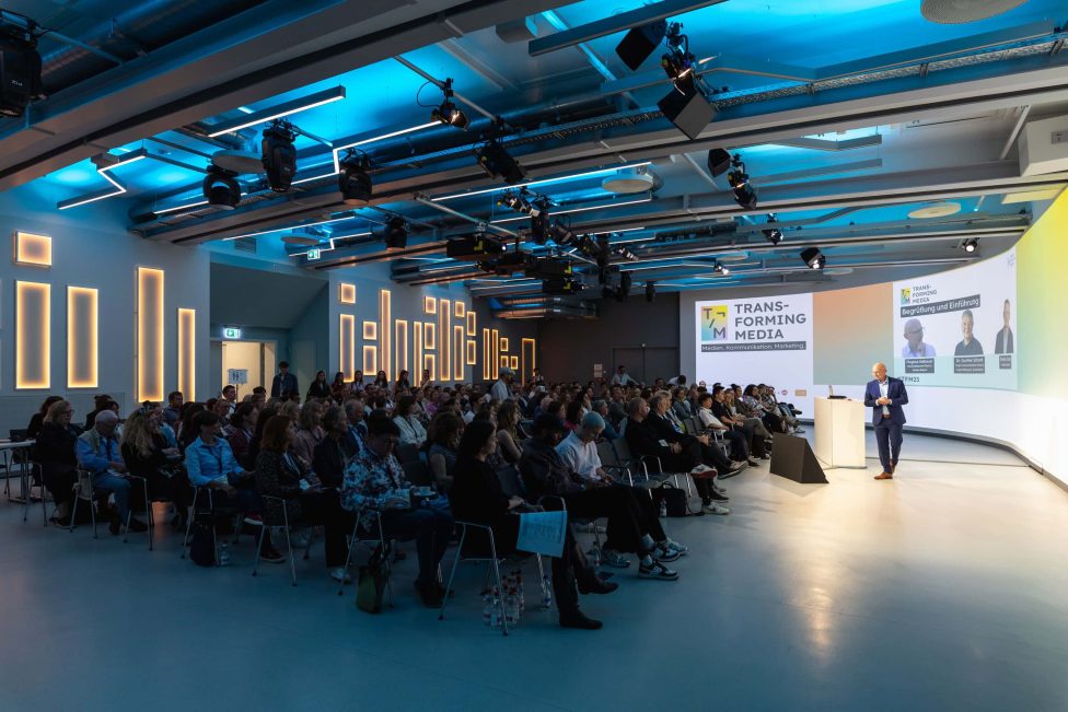 Ein vollbesetzter Konferenzraum mit Zuhörern, die einem Vortragenden auf der Bühne folgen. Die Veranstaltung 'Transforming Media 2025' wird auf einer großen Leinwand präsentiert. Die Beleuchtung ist modern und blau akzentuiert.