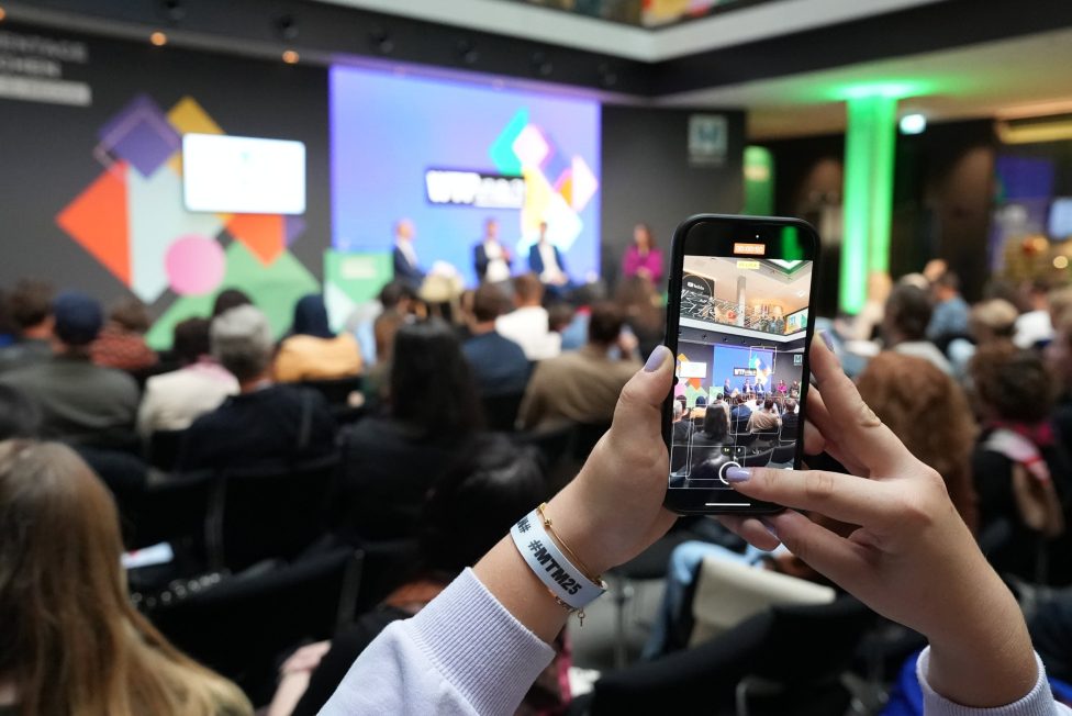 Das Bild zeigt eine Konferenz bei den MEDIENTAGEN MÜNCHEN 2025 im House of Communication. Im Vordergrund wird die Veranstaltung mit einem Smartphone gefilmt, während im Hintergrund eine Podiumsdiskussion vor einem farbenfrohen Bühnenbild stattfindet.