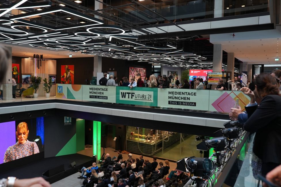 Das Bild zeigt die Medientage München 2025 im modernen House of Communication. Im Vordergrund eine Bühne mit Präsentation, im Hintergrund Networking-Bereiche und Besucher. Auffällig sind die futuristische Deckenbeleuchtung und die farbenfrohen Banner mit dem Motto 'WTFuture?!'.