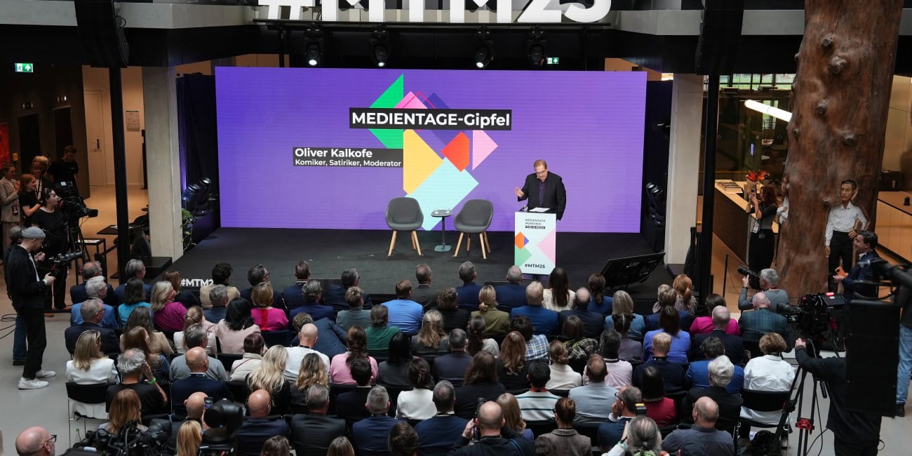 Auf der Bühne des MEDIENTAGE-Gipfels 2025 im House of Communication in München steht Oliver Kalkofe, Komiker, Satiriker und Moderator. Vor einer großen, aufmerksam lauschenden Zuschauermenge spricht er vor einer farbenfrohen Leinwand mit dem Hashtag #MTM25 im Hintergrund.