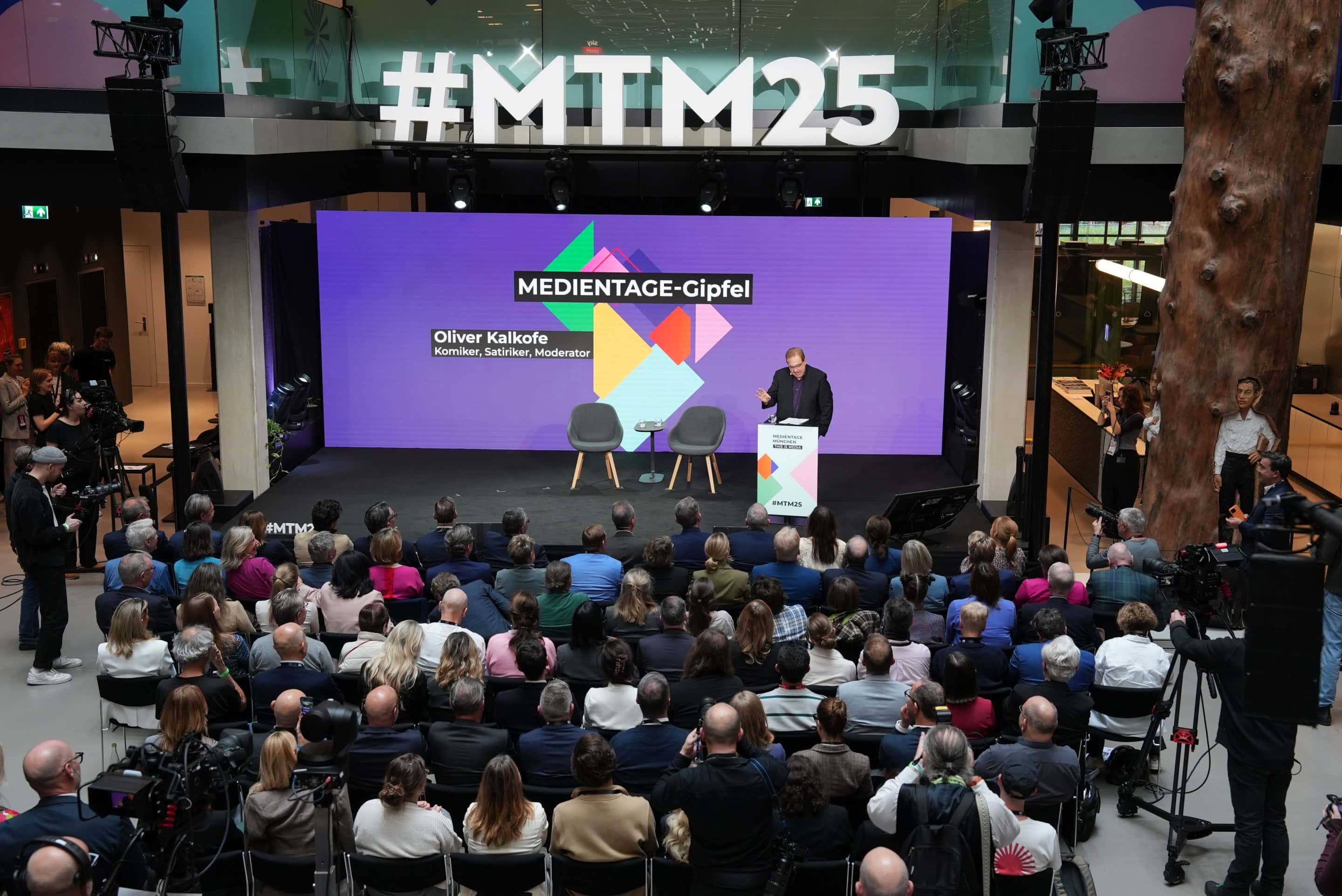 Auf der Bühne des MEDIENTAGE-Gipfels 2025 im House of Communication in München steht Oliver Kalkofe, Komiker, Satiriker und Moderator. Vor einer großen, aufmerksam lauschenden Zuschauermenge spricht er vor einer farbenfrohen Leinwand mit dem Hashtag #MTM25 im Hintergrund.