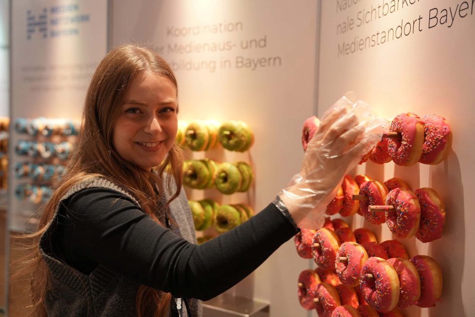 Eine Person arrangiert pink glasierte Donuts mit bunten Streuseln auf einer dekorativen Wand. Im Hintergrund sind weitere Donuts in verschiedenen Farben zu sehen. Die Szene zeigt eine kreative und einladende Atmosphäre auf der Konferenz.