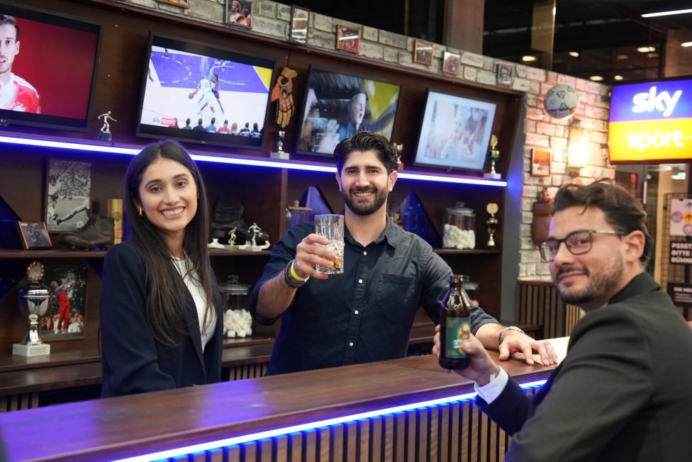 An einer stilvollen Bar mit beleuchteten Regalen und Sportübertragungen im Hintergrund stoßen Gäste mit Getränken an. Die Umgebung ist geprägt von sportlichen Trophäen und einer lockeren, kommunikativen Stimmung.