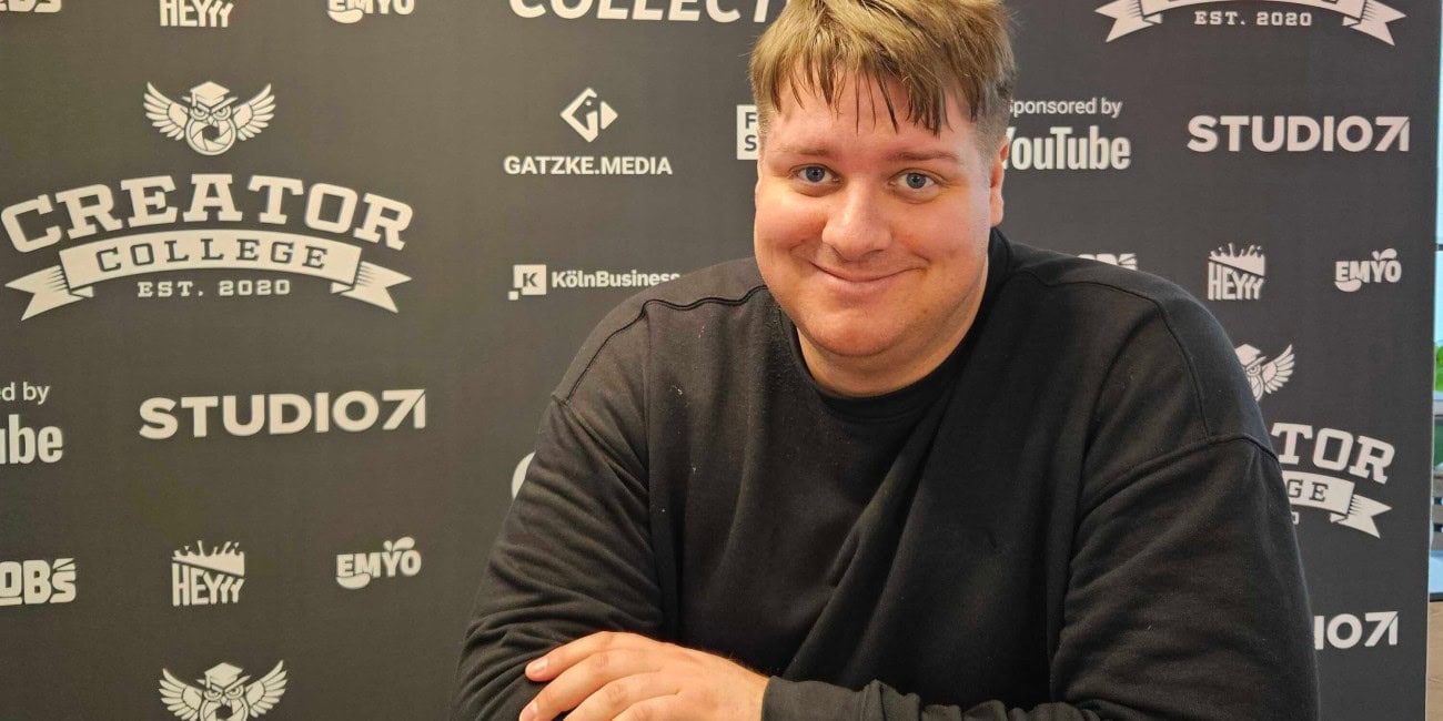 A manEin Mann sitzt vor einer Wand mit einem Schild, auf dem „Creator Collect“ steht.