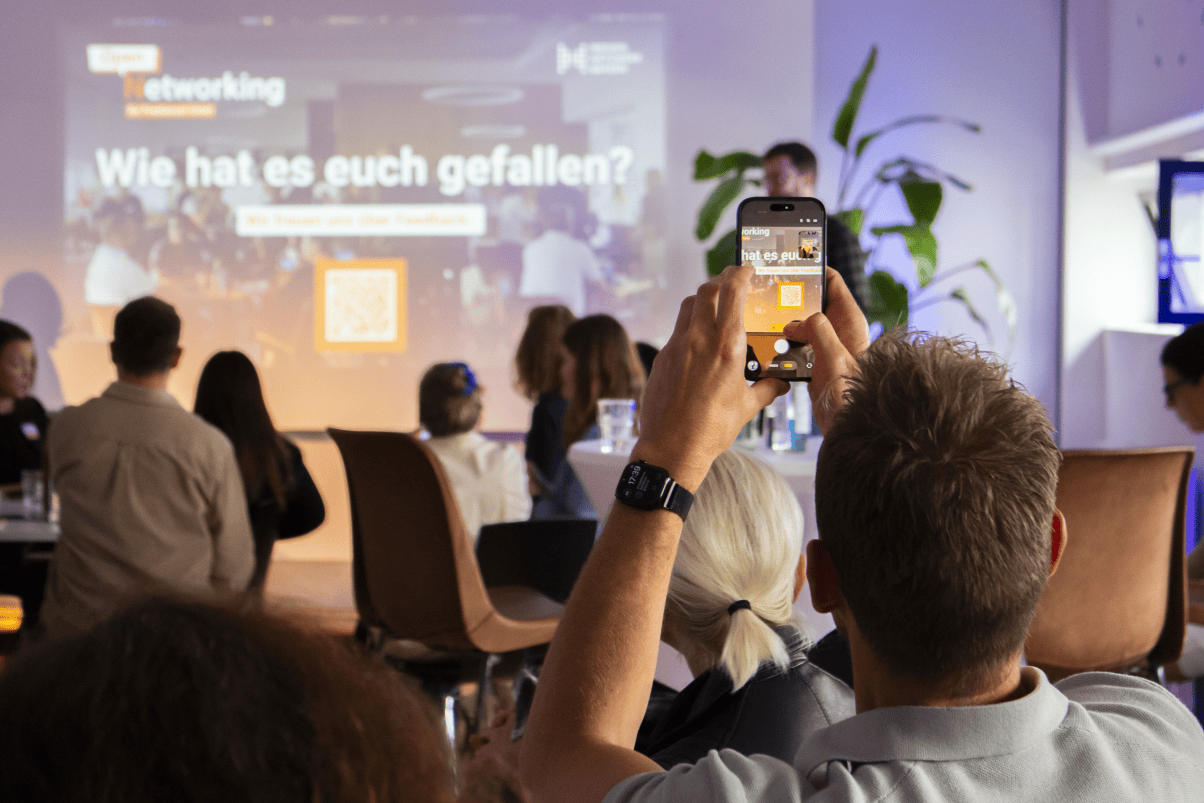 MNB Open Networking Menschen sitzen an Tischen in einem Veranstaltungsraum. Im Vordergrund hält eine Person ein Smartphone hoch, um die Präsentation mit einem QR-Code auf der Leinwand zu fotografieren. Pflanzen und moderne Einrichtung sind sichtbar.
