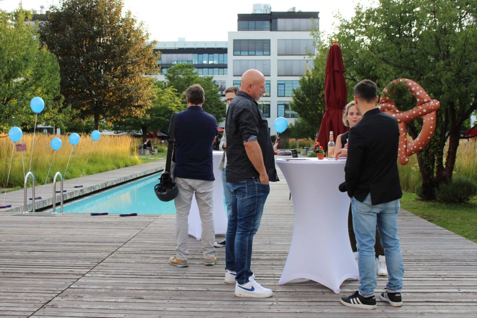 Mehrere Personen stehen an Stehtischen neben einem Pool im Freien. Im Hintergrund sind Bäume, ein Gebäude und blaue Luftballons zu sehen. Eine große Brezel-Dekoration ist ebenfalls sichtbar.