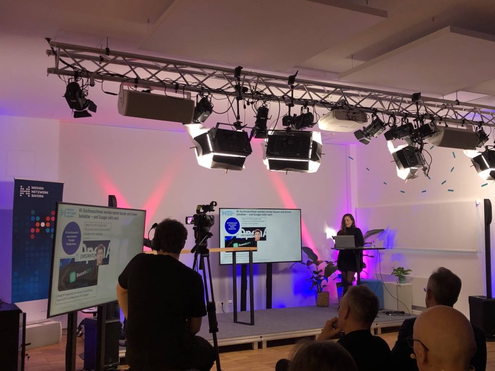 Das Bild zeigt eine Präsentation bei der Veranstaltung Medientrends 2025. Eine Sprecherin steht auf der Bühne vor einem Publikum, umgeben von Kameras und Bildschirmen. Die Beleuchtung ist professionell eingerichtet.