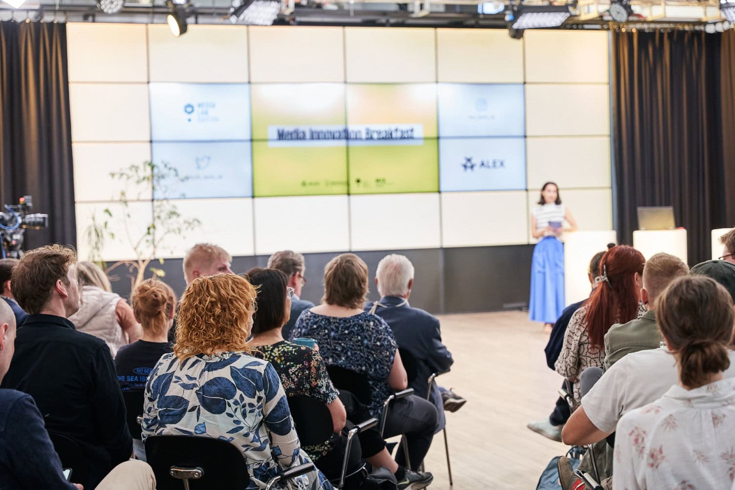 Media Innovation Event im Media Lab Bayern Das Bild zeigt eine Gruppe von Menschen, die in einem Veranstaltungsraum sitzen und einer Präsentation folgen. Im Hintergrund ist eine große Leinwand mit dem Titel 'Media Innovation Broadcast' zu sehen. Eine Frau steht vorne und hält eine Rede.