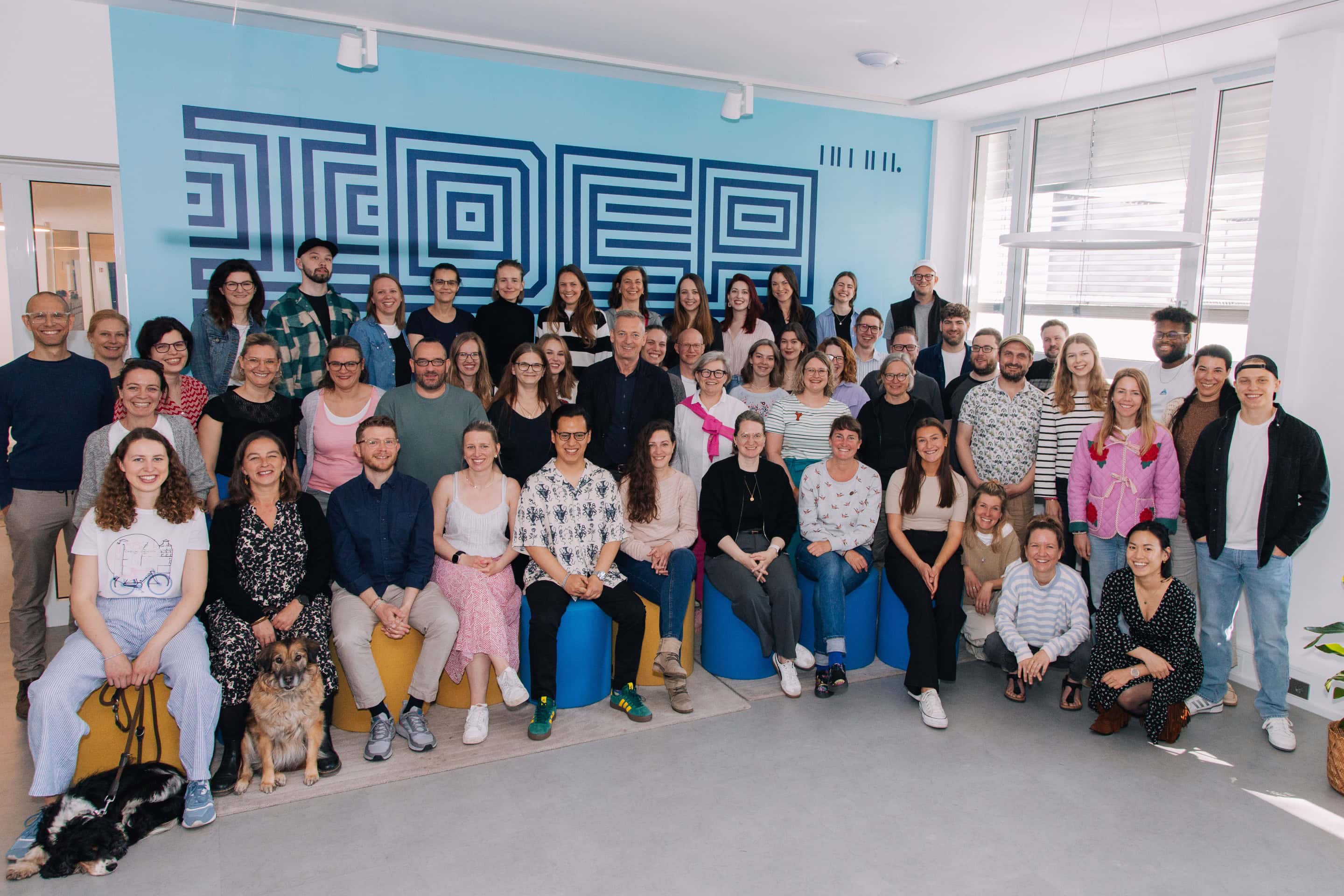 Die Mitarbeiterinnen und Mitarbeiter der Medien.Bayern und ihre beiden Geschäftsführer Stefan Sutor und Annette Kümmel stehen lachend vor der Ideas Café Wand im Büro der Medien Bayern GmbH in München