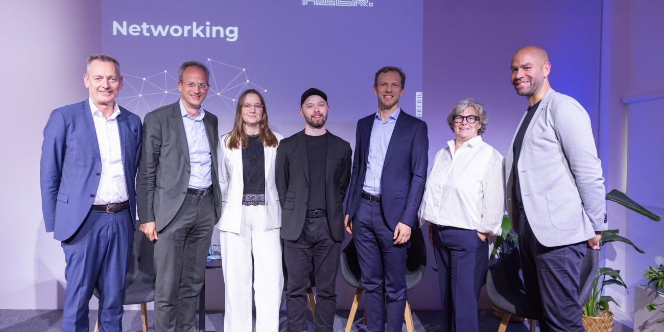 Sieben Personen in Business-Outfits stehen nebeneinander auf einer Bühne. Dahinter eine violette Leinwand mit Logos „Medien.Bayern“, „MedienNetzwerk Bayern“ und Schriftzug „Zukunft beginnt hier – Networking“. Seitlich Zimmerpflanzen.