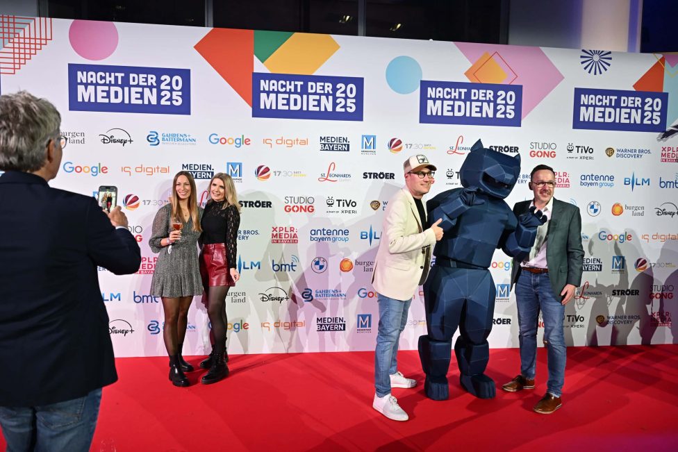 Auf dem roten Teppich der 'Nacht der Medien 2025' posieren Gäste vor einer Sponsorenwand. Ein auffälliges Maskottchen in Blau sorgt für Unterhaltung, während ein Fotograf die Szene festhält. Die Veranstaltung bietet eine lebendige Atmosphäre für Medienschaffende.