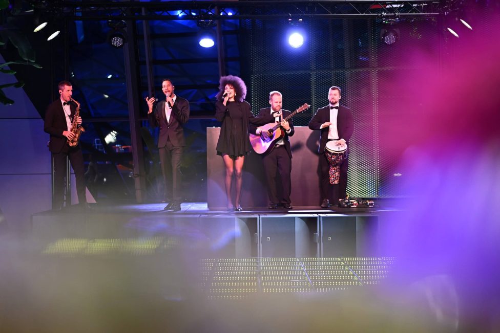 Eine stilvolle Band in eleganter Abendgarderobe performt auf einer Bühne mit stimmungsvoller Beleuchtung. Im Hintergrund moderne Architektur, im Vordergrund verschwommene Lichteffekte, die eine dynamische Atmosphäre schaffen.