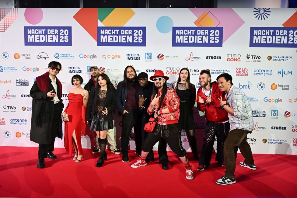 Eine Gruppe von Gästen posiert auf dem roten Teppich vor der Sponsorenwand der 'Nacht der Medien 2025'. Die Teilnehmer präsentieren sich in stilvollen und individuellen Outfits, die von elegant bis urban reichen. Die Atmosphäre ist lebendig und kreativ.