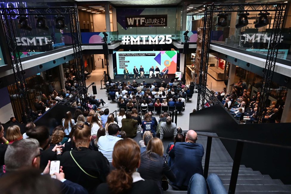 Das Bild zeigt eine Podiumsdiskussion während der MEDIENTAGE MÜNCHEN 2025 im House of Communication. Vor einer großen Leinwand diskutieren Experten auf einer Bühne, während zahlreiche Gäste auf Sitzplätzen und Treppen aufmerksam zuhören. Die Veranstaltung ist gut besucht und professionell organisiert.