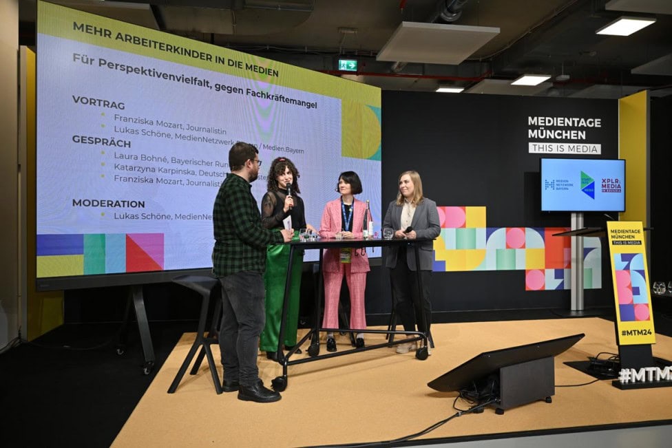 Das Bild zeigt ein Panel auf den Medientagen München mit vier Personen, die an einem Stehtisch diskutieren. Im Hintergrund ist eine große Leinwand mit dem Thema 'Mehr Arbeiterkinder in die Medien' und Logos der Veranstalter zu sehen.