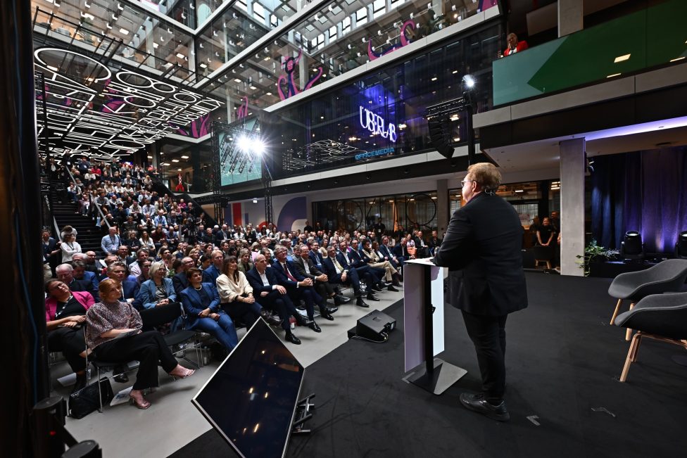 Ein Redner spricht vor einem großen Publikum im modernen Ambiente des House of Communication in München. Die Veranstaltung ist geprägt von einer dynamischen Atmosphäre, mit einer beeindruckenden Glasarchitektur und aufmerksamem Publikum.