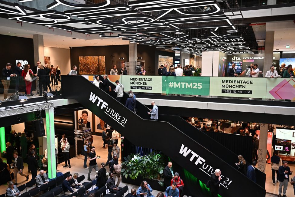 Das Bild zeigt die belebte Atmosphäre der Medientage München 2025 im House of Communication. Besucher tauschen sich aus, während moderne Architektur, kreative Lichtinstallationen und der Schriftzug 'WTFuture 21' auf der Rolltreppe das innovative Ambiente unterstreichen.