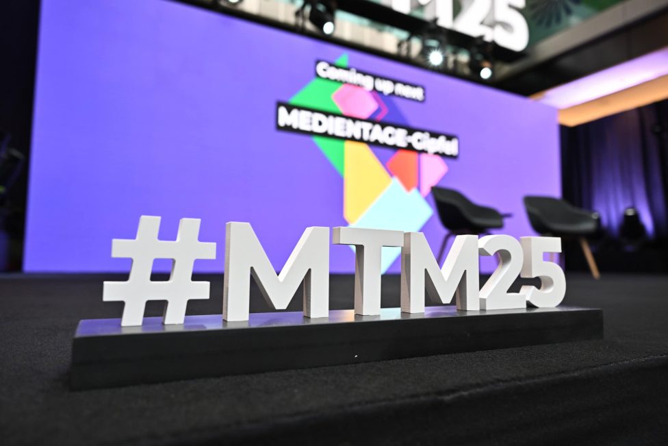 Das Bild zeigt eine Bühne der MEDIENTAGE MÜNCHEN 2025 im House of Communication. Im Vordergrund steht ein dekoratives Hashtag #MTM25, während im Hintergrund eine bunte Leinwand mit der Aufschrift 'Coming up next MEDIENTAGE-Club' und zwei leere schwarze Stühle zu sehen sind.
