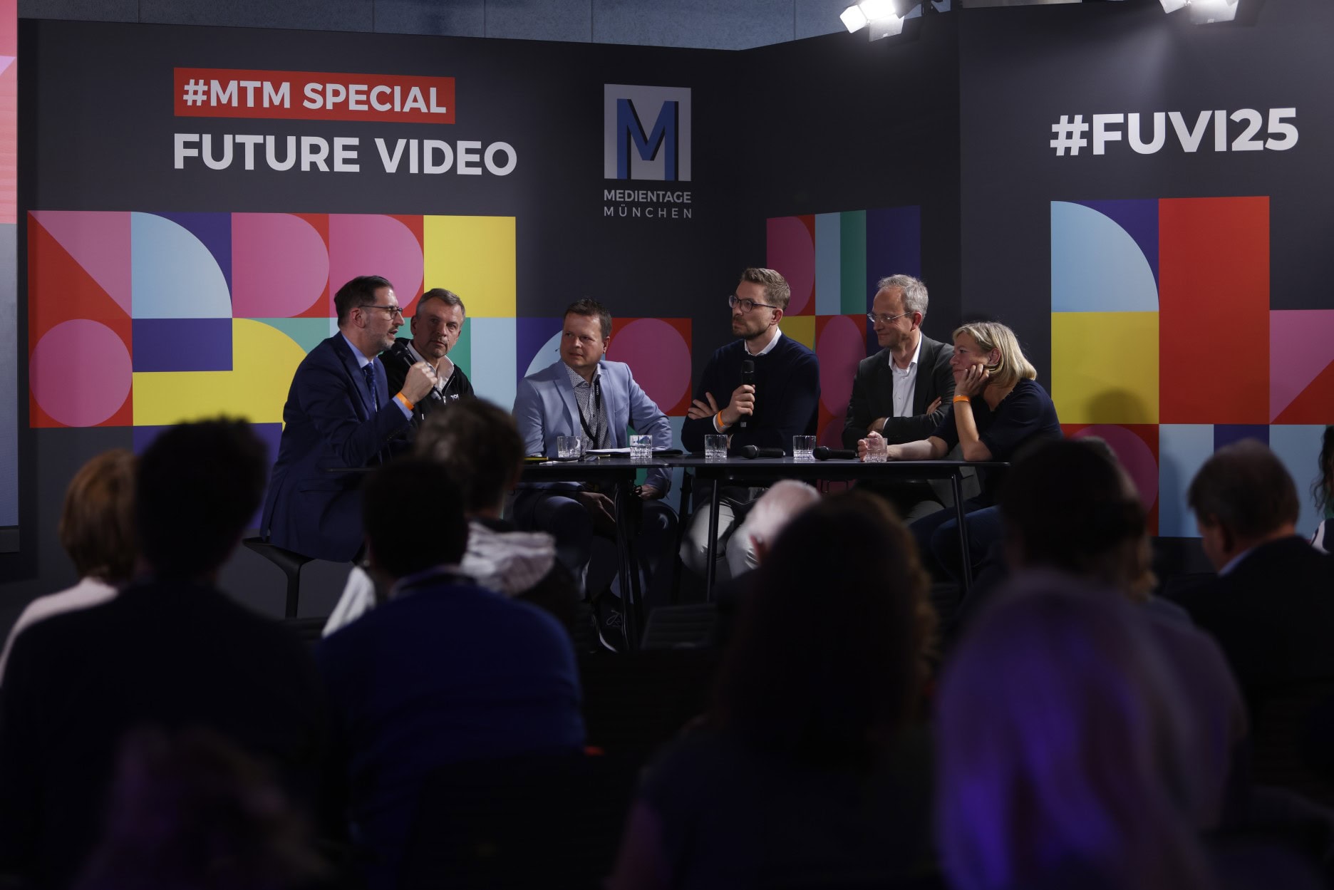 Eine Gruppe von sechs Personen sitzt an einem Tisch und diskutiert auf einer Bühne. Im Hintergrund sind bunte geometrische Muster und die Schriftzüge '#MTM SPECIAL Future Video' und '#FUVI25' zu sehen.