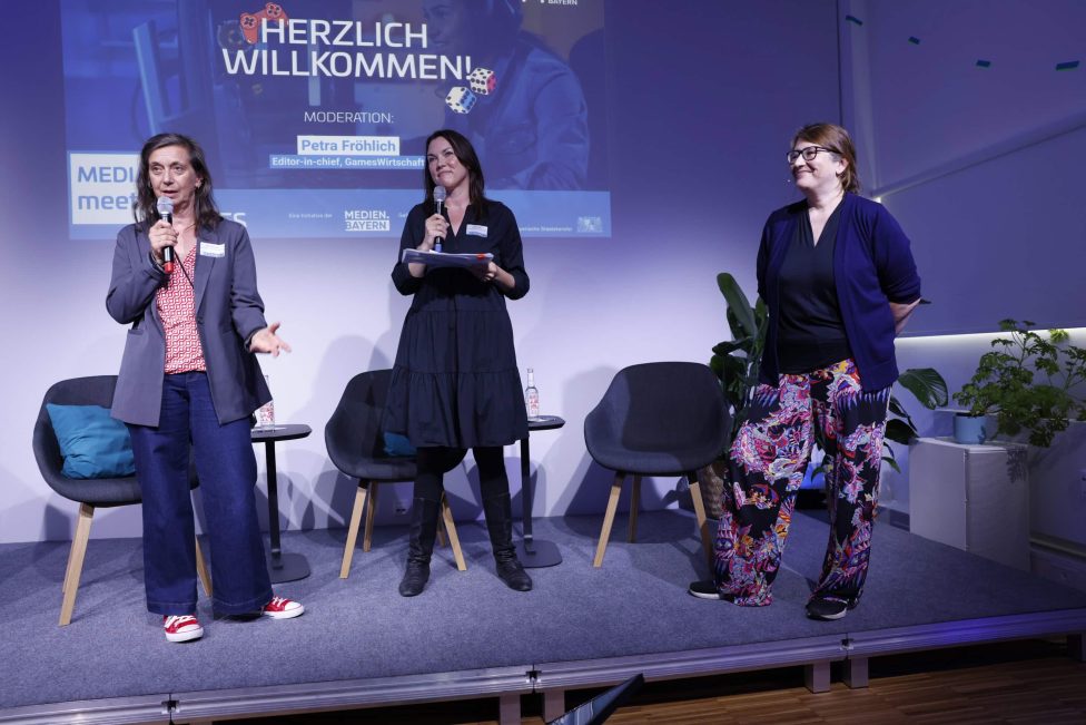Drei Frauen stehen auf einer Bühne vor einem projizierten Begrüßungsscreen. Zwei halten Mikrofone, eine trägt bunte Kleidung. Im Hintergrund sind Pflanzen und Sitzgelegenheiten zu sehen.
