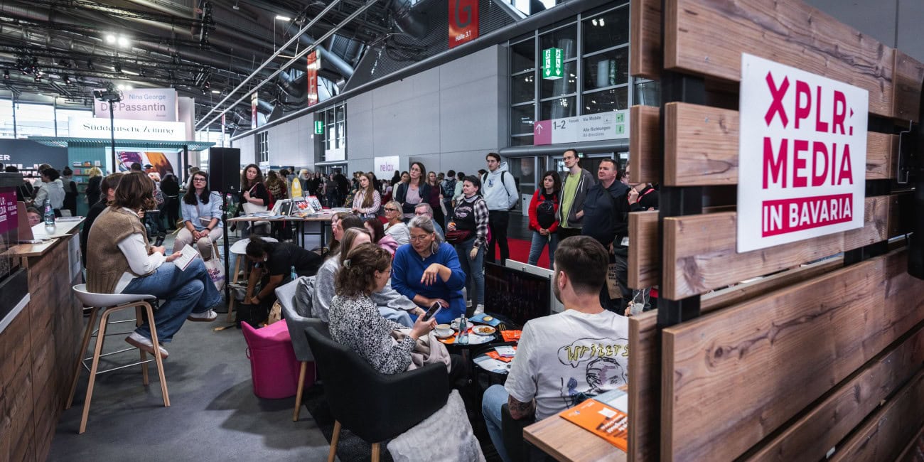 Am Stand von XPLR: MEDIA in Bavaria sitzen Menschen in einer gemütlichen Lounge-Atmosphäre, umgeben von Büchern und Informationsmaterial. Im Hintergrund sind weitere Messestände und Besucher der Frankfurter Buchmesse zu sehen.