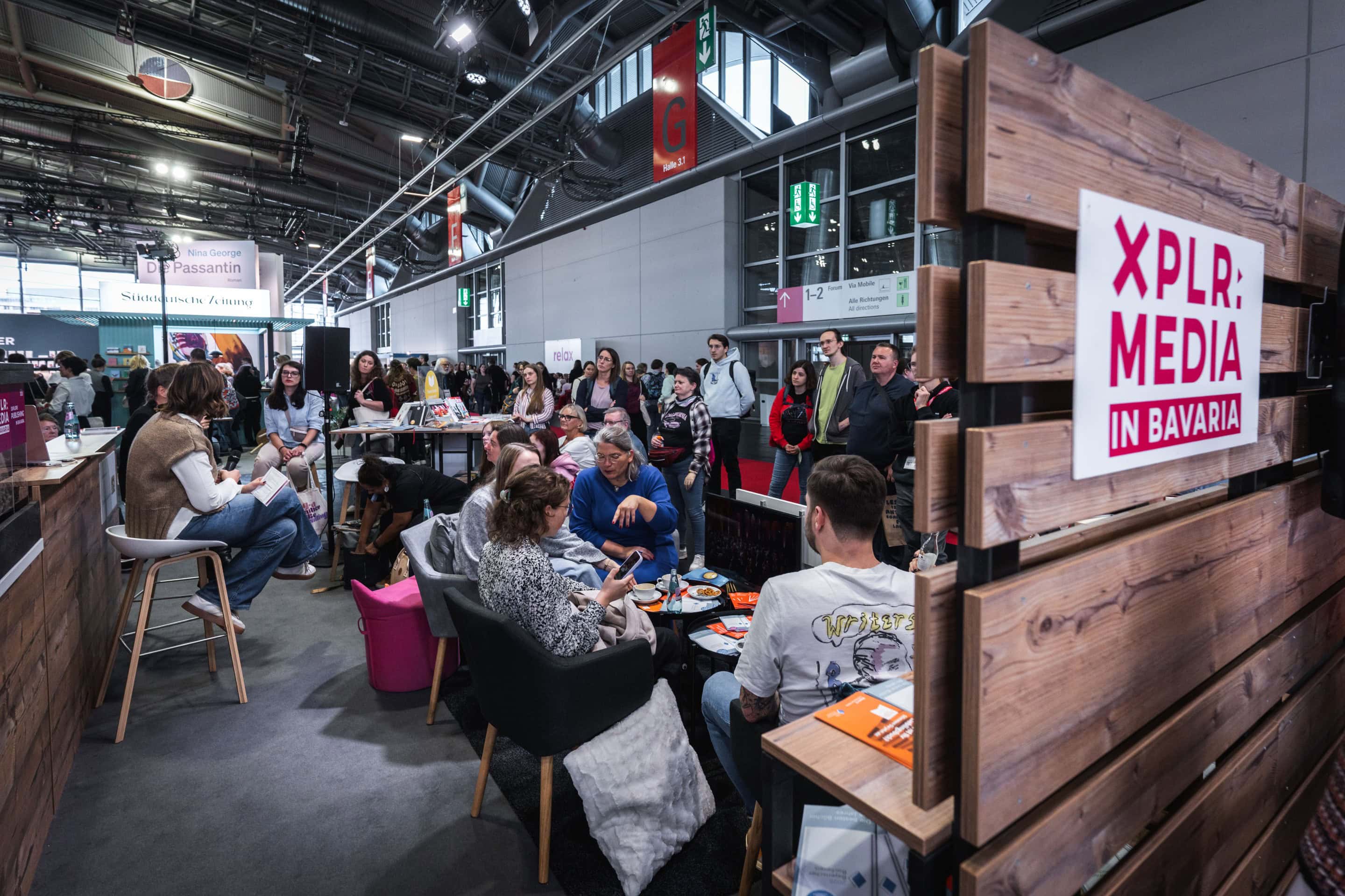 Am Stand von XPLR: MEDIA in Bavaria sitzen Menschen in einer gemütlichen Lounge-Atmosphäre, umgeben von Büchern und Informationsmaterial. Im Hintergrund sind weitere Messestände und Besucher der Frankfurter Buchmesse zu sehen.